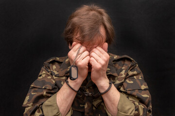 A military man in handcuffs covers his face, holds a military badge in his hand on a dark background, selective focus.