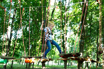 Small boy in climbing gear is walking along a rope road in an adventure Park, holding on to a rope and a carabiner.