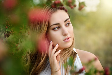 A beautiful young girl with long brown hair and brown eyes against a background of a green tree with bright red berries. Autumn portrait of a girl with professional makeup