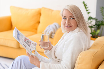 Mature woman with glass of water reading newspaper in armchair at home