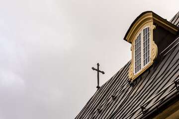 Fototapeta na wymiar Metal cross at the rooftop against grey cloudy sky at the roof of the building. Detail of the church roof with cross and decorated roof window. Cross protecting the sky on roof.