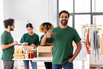 charity, donation and volunteering concept - happy smiling male volunteer pointing to himself and international group of people packing food in boxes at distribution or refugee assistance center