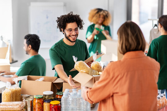 charity, donation and volunteering concept - happy smiling male volunteer with clipboard and woman taking box of food at distribution or refugee assistance center