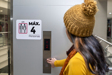 Unrecognizable young Latin American woman (23) in a shopping mall presses the elevator button. She wears a long-sleeved flannel and a yellow cap. Concept of moving