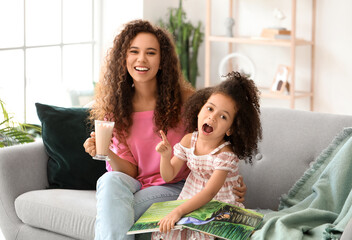 Little African-American girl and her mother with glass of hot cocoa and book at home
