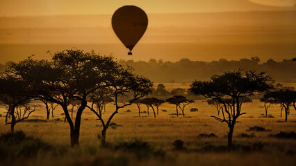 Bright sunset on a field with a hot air balloon, trees, and animals in Masai Mara, Kenya