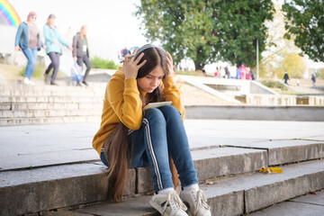 A sad teenage girl sits on the stairs and covers her head with her hands