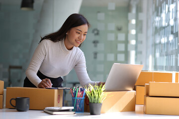 Starting a Small Business SME Owner Female Entrepreneur Reading and writing order numbers. Receiving and reviewing online orders to prepare to pack boxes, sell to customers, sme business ideas online. - obrazy, fototapety, plakaty