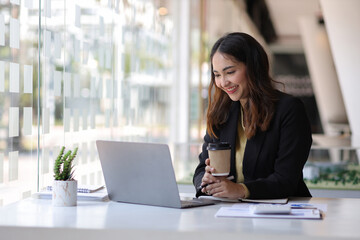 Attractive Asian secretary or businesswoman holding hot coffee mug sitting on laptop in office.