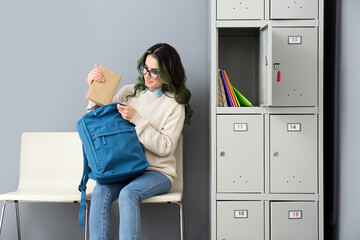 Beautiful female student putting book in backpack while sitting near locker at the university - Powered by Adobe