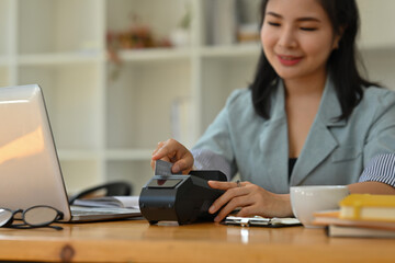 Cropped view of a smiling Asian woman sitting in the office holding a credit card and using a credit card swipe machine, for finance and technology concept.