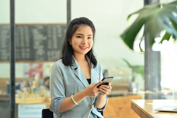 A portrait of a pretty Asian woman sitting in the office using a smartphone and looking at the camera with confidence, for business and technology concept.