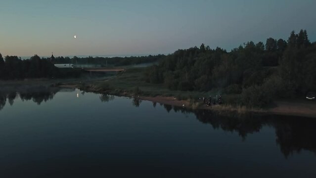 Mist over the river in the early morning dawn. A group of young people resting and singing songs on the shore, woods and countryside in the background 