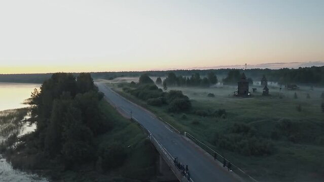 Young people on a lonely road on a bridge near a church on a foggy morning at dawn. River, forest, fog, village