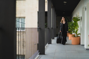 Chica joven con pelo largo vestida de negro con maleta de viaje con ruedas por pasillo de hotel...