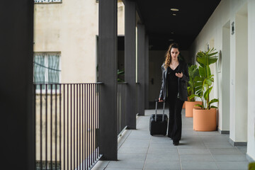 Chica joven con pelo largo vestida de negro con maleta de viaje con ruedas por pasillo de hotel...