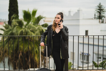 Chica joven con pelo largo y vestida de negro con maleta de viaje en azotea de un hotel estilo soho