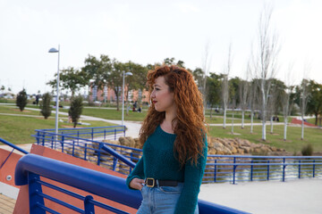 Obraz na płótnie Canvas pretty young red-haired woman is leaning on a railing in a park. She is posing for the photo smiling. Fashion and beauty concept
