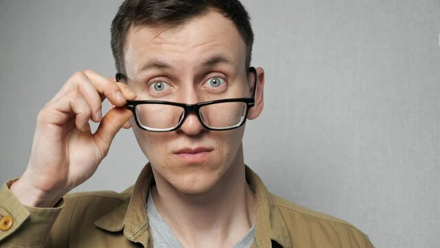 Man with bad eyesight removes black-rimmed glasses getting surprised. Emotional male person shows astonishment on grey background close view