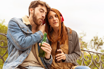 Young couple listening music on one headphones with beer bottles in hand - sitting on bench in the...