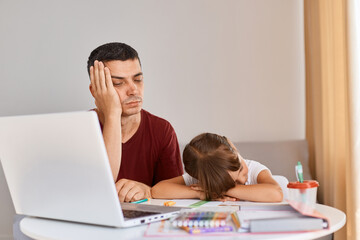 Indoor shot of tired sleepy young adult father sitting with schoolgirl daughter at table, family posing with closed eyes, being exhausted to do hard tasks.