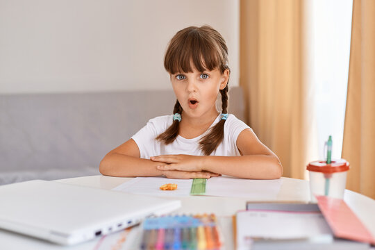 Image of astonished charming child girl doing homework reading shocking information, posing at home, looking at camera with widely open mouth, distance education.