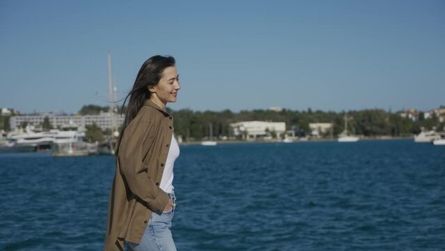 A happy girl, a brunette, of Asian appearance, walks along the promenade on the seashore, smiles and looks at the camera. The wind blows hair, the sun shines. Medium plan.
