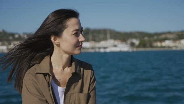 A happy girl, a brunette, of Asian appearance, sits on a pier by the sea, smiles. The wind blows your hair, the sun shines. Close-up.