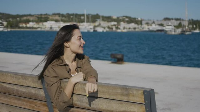A happy girl, a brunette, of Asian appearance, sits on a bench, on a pier by the sea, smiles and looks into the camera. The wind blows your hair, the sun shines. Medium plan.