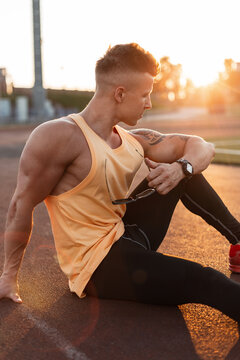 Handsome young stylish guy with a healthy athletic body in a yellow tank top, black leggings and sneakers sits on the street at sunset