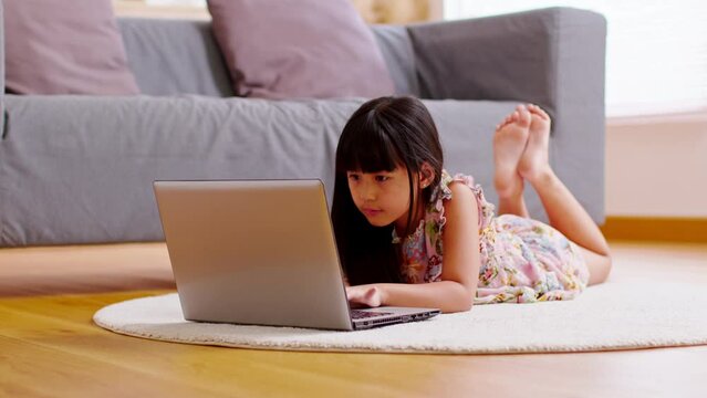Cute little girl uses laptop while prone at the sofa in the living room. Child surfing the internet on computer, browses through internet and watches cartoons online.