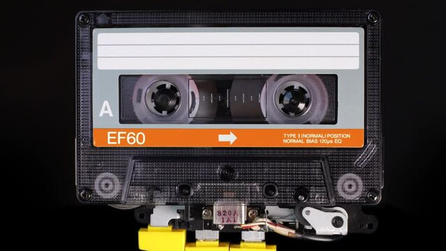 A vintage, orange colored compact cassette tape playing back in a player mechanism. Tape recorder head and rotating reels of deck player. Magnetic tape stores stereo music in a magnetic image
