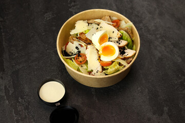 Caesar salad with chicken meat in paper take away container with anchovy sauce isolated on black background. Healthy food concept, restaurant dish delivery. Top view. Free space for your text.