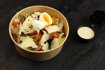Caesar salad with chicken meat in paper take away container with anchovy sauce isolated on black background. Healthy food concept, restaurant dish delivery. Top view. Free space for your text.