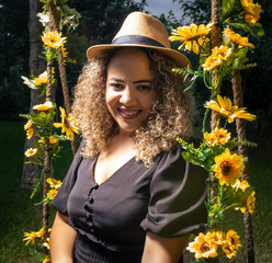 Brown woman with curly hair and black dress with nude hat looking forward surrounded by yellow flowers and green leaves with a deep gaze towards the light. Mention of female empowerment.