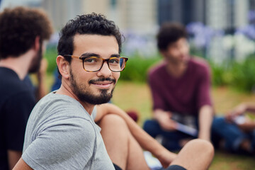 Loving life on campus. Portrait of a cheerful young man sitting in the park with his friends and studying outside during the day.