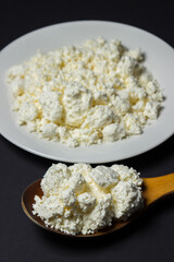 A plate of cottage cheese on a black background. Healthy and tasty dairy product. Wooden spoon full of cottage cheese