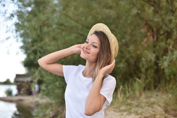 young woman in white t-shirt holds a straw hat with her hands and looks at the blue sky sitting on the shore. girl dreaming outdoors