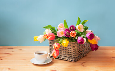Obraz na płótnie Canvas Colorful fresh tulips in wicker basket and cup of coffee on wooden table. Selective focus.
