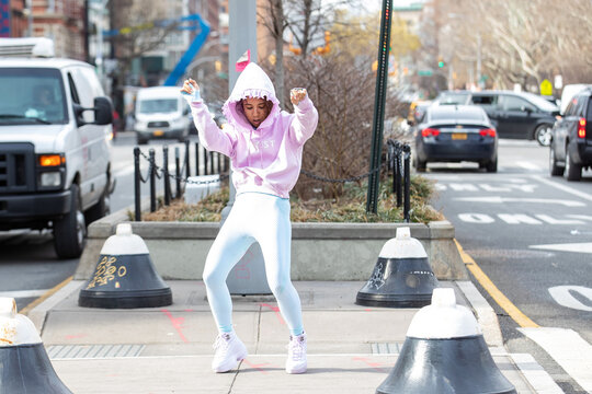 Woman wearing pink hoodie with the word artist dances outside in New York city street
