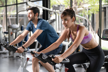 Fototapeta na wymiar Young sporty couple doing cardio work out on stationery exercise bikes in indoor fitness gym. Man and woman exercising and cycling bikes together in gym. Healthy lifestyle concept.