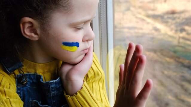 Portrait of unhappy sad little girl child with flag of ukraine on face sitting alone on windowsill at home. Protest against russian war invasion in Ukraine. Ukrainian Flag, Protesters concept
