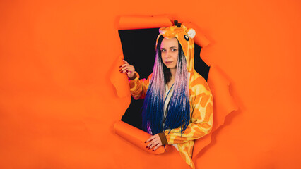 Young woman in pajamas of giraffe in hole of orange background. Pretty female in kigurumi looking at camera and smiling.