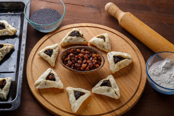 Gomentash with poppy seeds and prunes freshly baked for the holiday of Purim on a round board next to poppy seeds and raisins and a rolling pin.