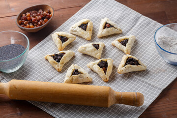 Gomentashash with poppy seeds and prunes freshly baked for the Purim holiday on a gray checkered towel next to a rolling pin and poppy seeds.