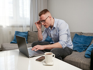 A young man with glasses sits in his room on the sofa at a coffee table. He is shocked and looks at the laptop monitor in surprise. Next to it is a cup of coffee. Home office.