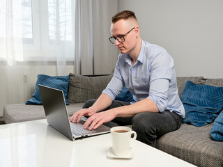 A young man with glasses sits in a room on a sofa at a coffee table and works on a laptop. Next to it is a cup of coffee. Home office.