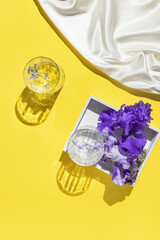 Two glasses with water on yellow background with white silk cloth and wooden tray with violet iris flowers. Summer refreshment concept. Top view. Flat lay.