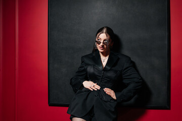 Attractive female posing in front of a black painting on a red wall wearing black jacket and...