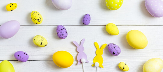 Easter eggs are yellow, lilac and purple, paper rabbits lie on a white wooden table in the form of a pattern. Copy space, flat lay